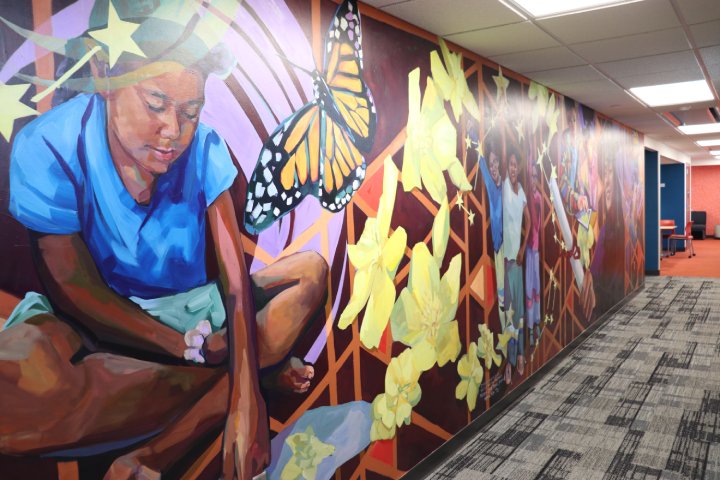 A vibrant mural on a hallway wall depicting a child sitting with a butterfly, surrounded by large yellow flowers and geometric patterns. The mural extends down the hall, featuring more children and colorful elements.